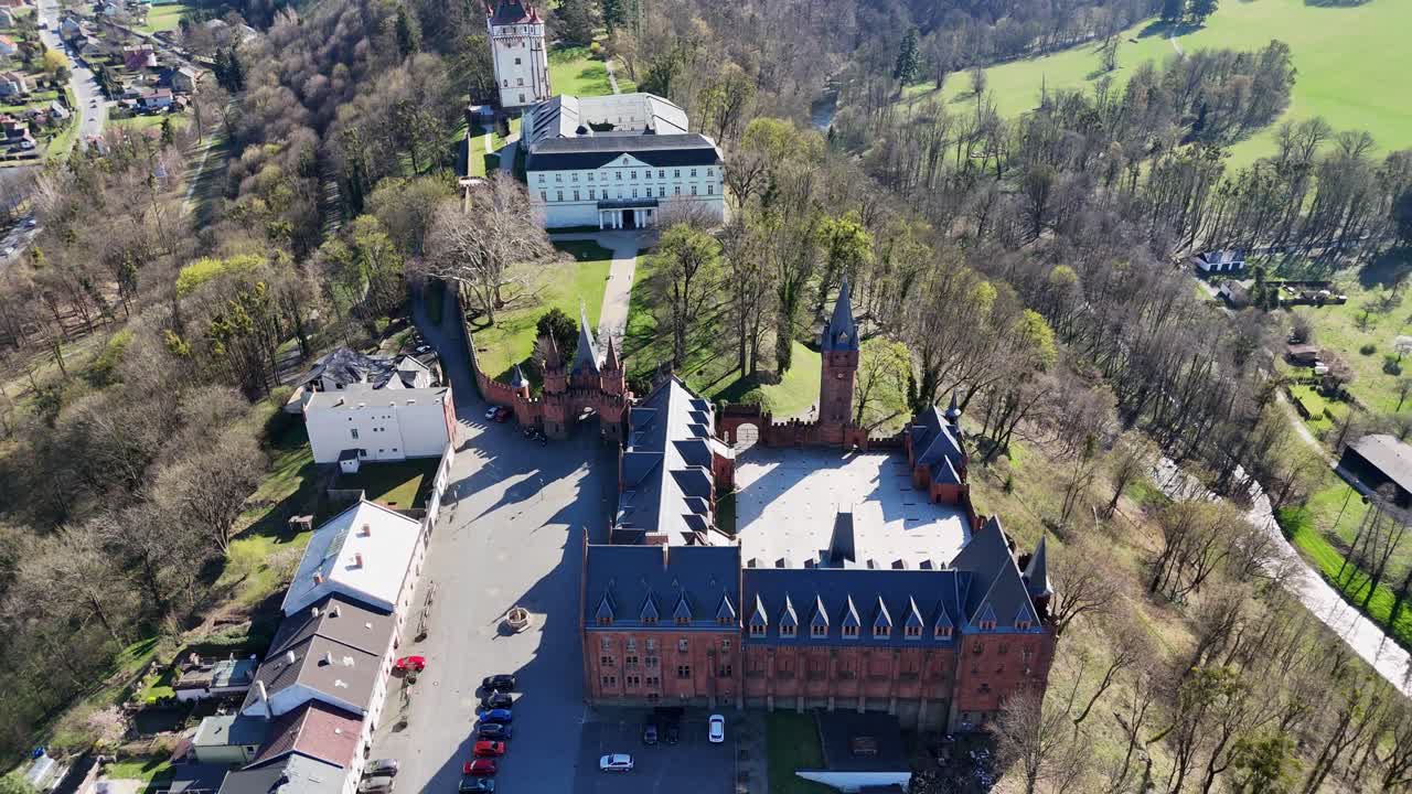 Zpětný let, kdy dron pomalu naklání kameru vzhůru, odhaluje zámecký komplex Hradec nad Moravicí v České republice s okolními kopci, vesnicí a širokou venkovskou krajinou