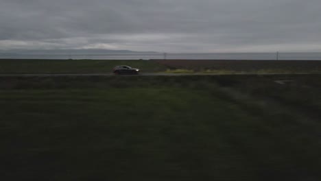 a car traverses a secluded road at dusk, its headlights cutting through the twilight as the imposing silhouette of distant mountains looms on the horizon