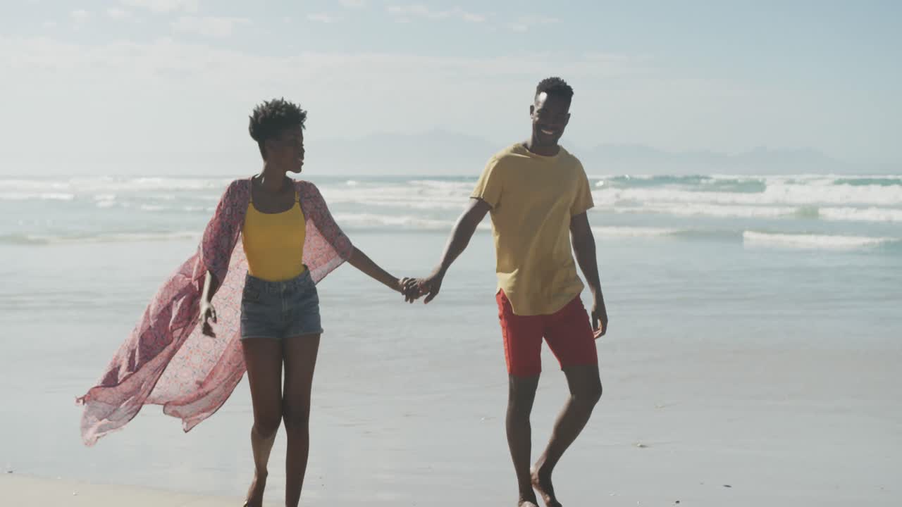 African american couple holding hands smiling while walking on the beach