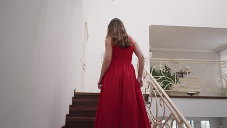 lady in red dress ascend stairs with elegance. young woman comes back to bedroom in private residence after exclusive party. tone for evening of glamour