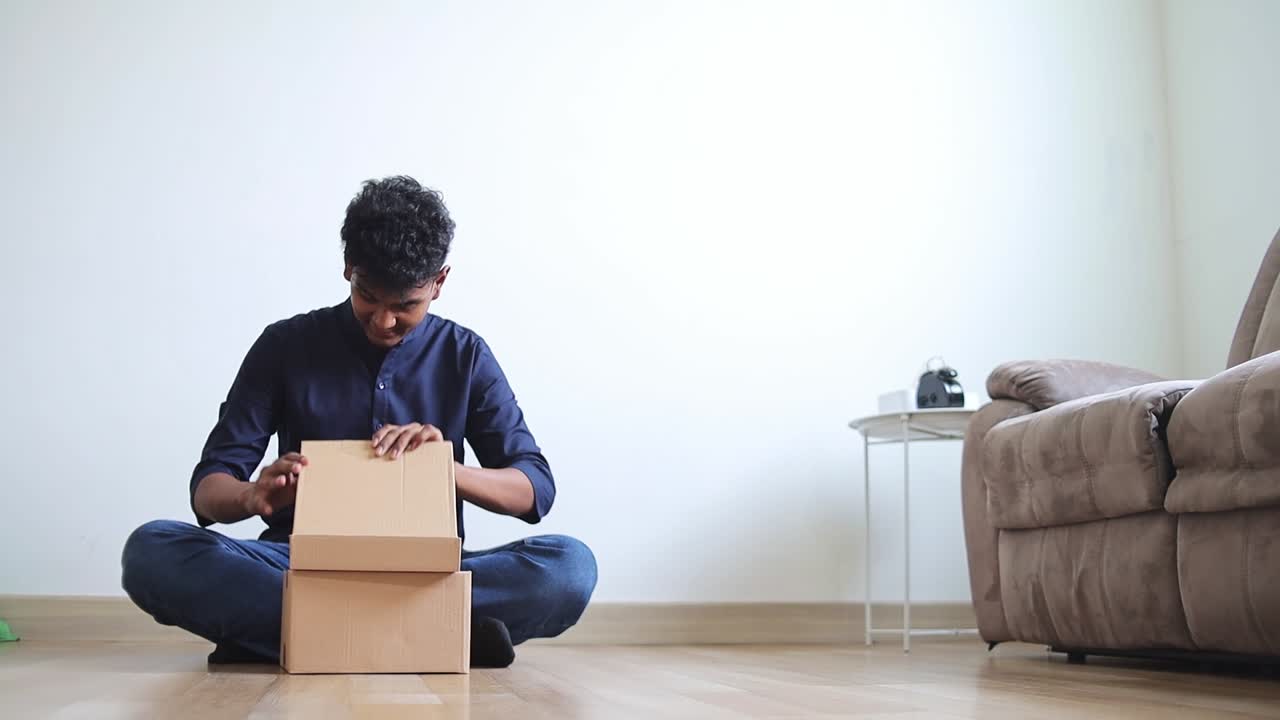 Excited young indian man unboxing package while receiving online order at modern home