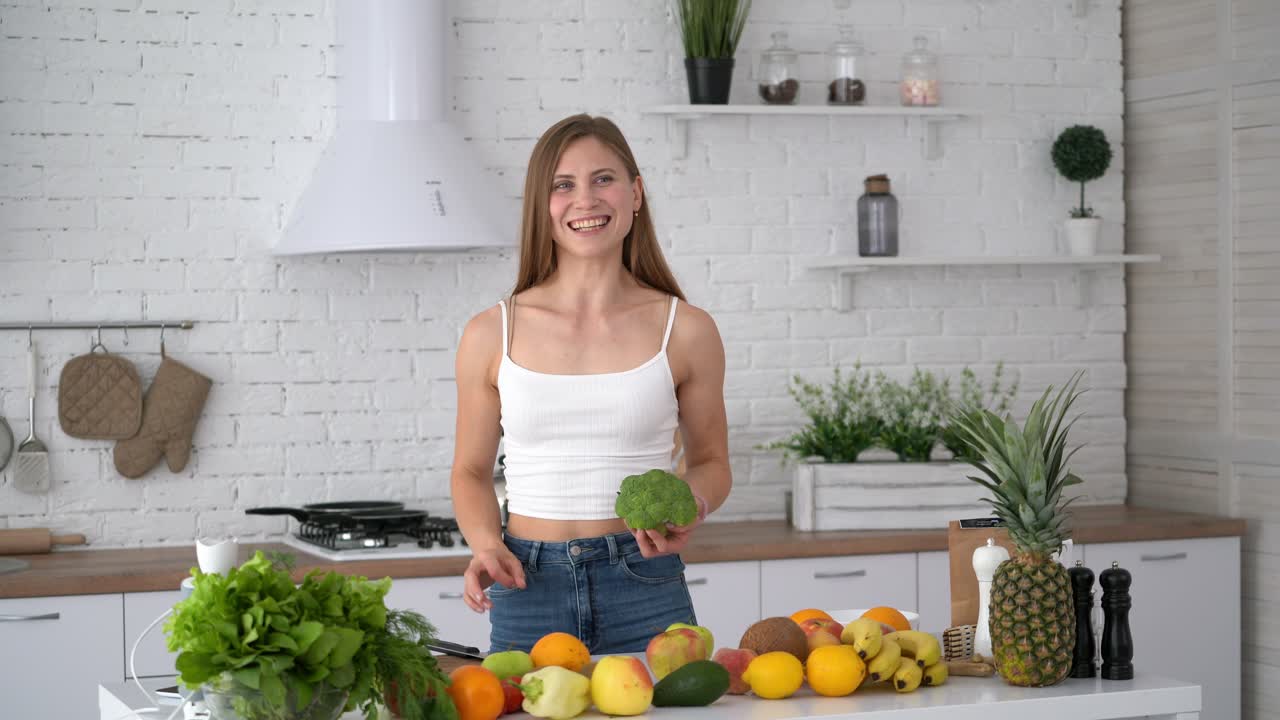 Healthy woman with sporty body in the kitchen. Attractive girl holding broccoli and shows her biceps on camera. Healthy food on the kitchen table at home.