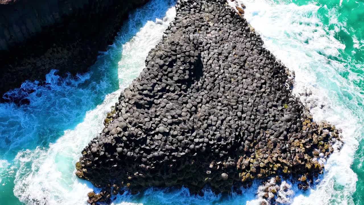 Aerial view of a triangular rock formation surrounded by vibrant turquoise ocean waves and lush greenery.