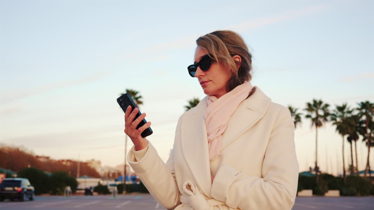 Donna elegante con cappotto bianco, sciarpa e occhiali da sole che controlla il telefono in una zona esterna soleggiata con palme