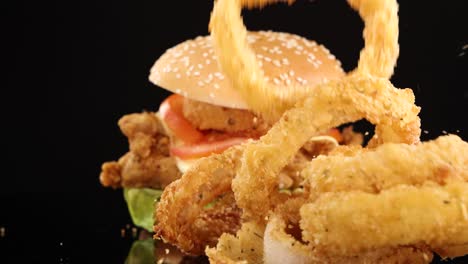 golden onion rings fall in slow motion next to a stacked fried chicken burger with lettuce, tomato, and cheese, shot against a black background with dramatic lighting