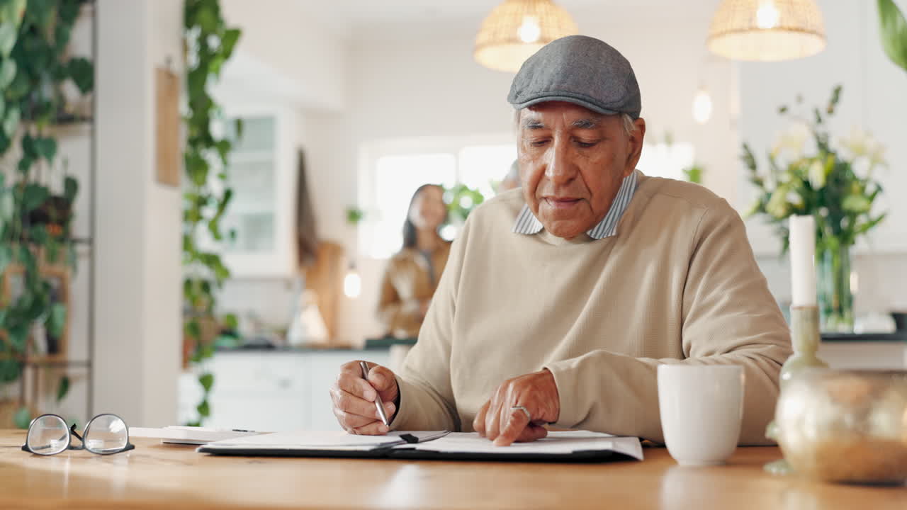un hombre mayor se sienta en una mesa en una cocina escribiendo en un cuaderno.