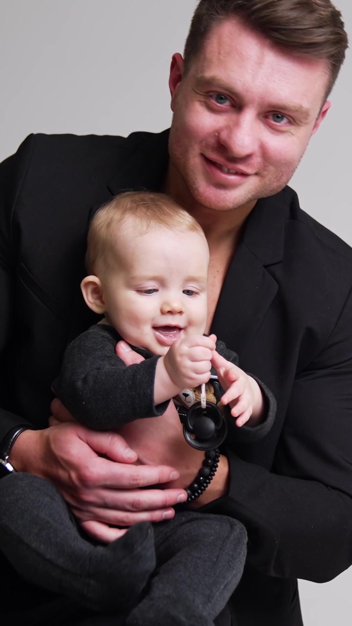 Caucasian man wearing black jacket holding a little cute baby in hands. Little kid is focused on the pacifier. Dad and son concept. White backdrop. Vertical video