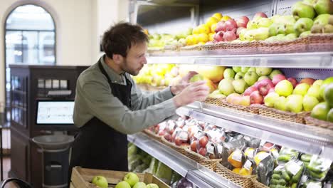 un vendedor ocupado está poniendo frutas en los estantes en la sección de verduras del supermercado , un tipo barbudo lleva un delantal