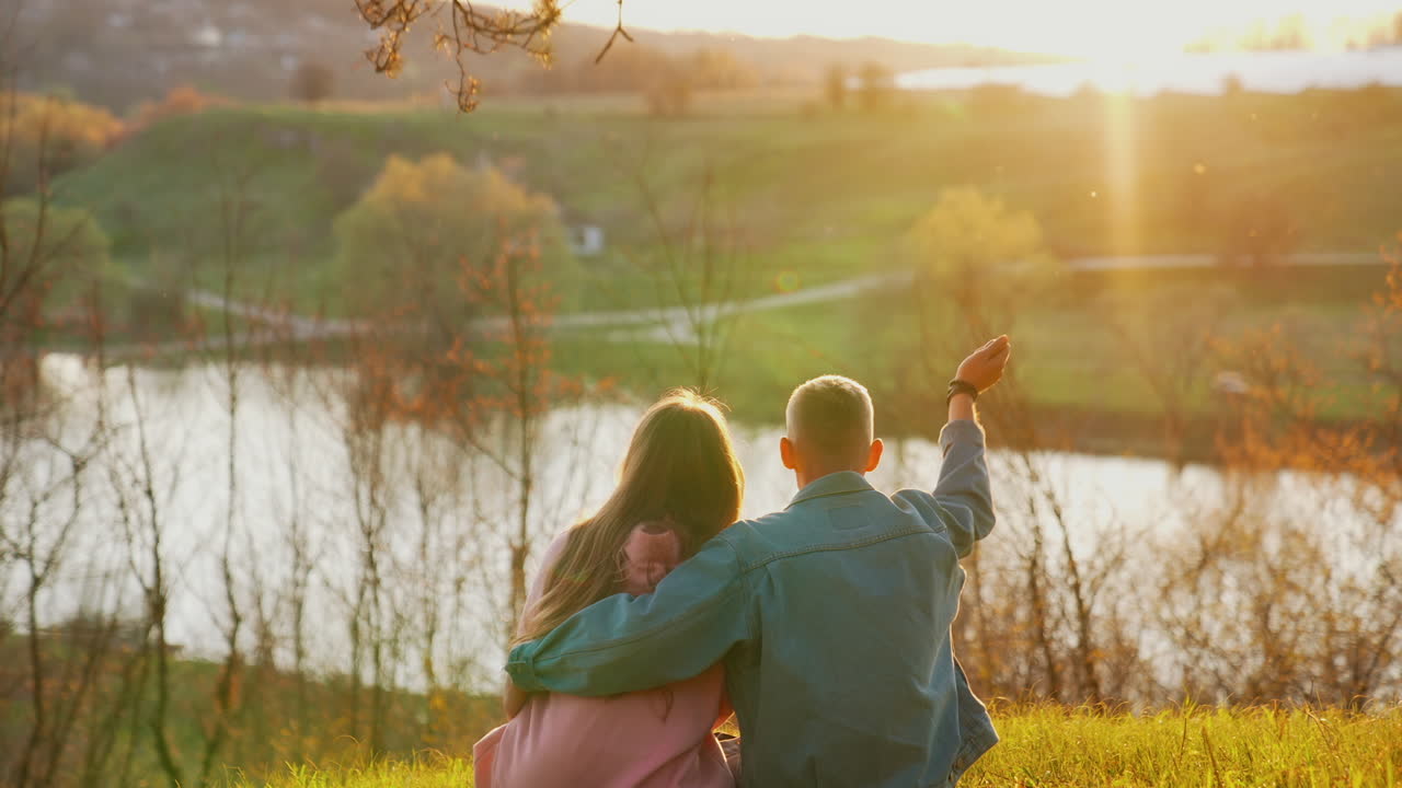 Young people sitting on grass and looking at sunset near the river. Guy embracing his girlfriend against autumn landscape in the evening.