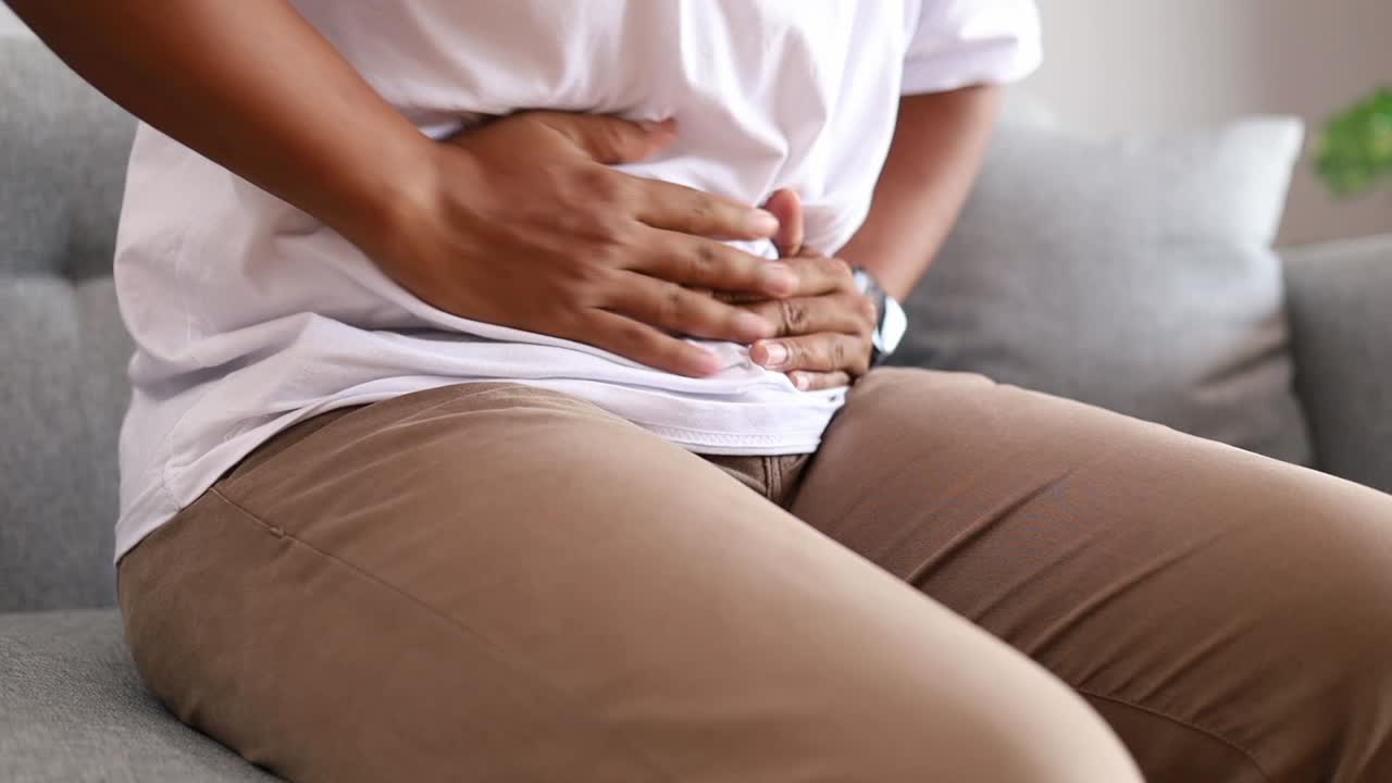Man's Hands holding stomach while sitting on sofa at home, tummy ache, indigestion, diarrhea or food poisoning