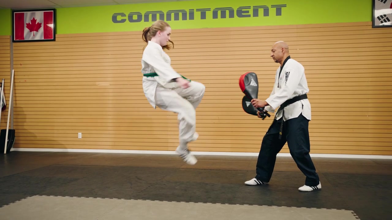 Ung kvinnelig kampsportutøver (Taekwondo, Karate) trener raske slag og spinnende dobbelt høyt spark mot slagpute med svartbelte-mester i treningshall.