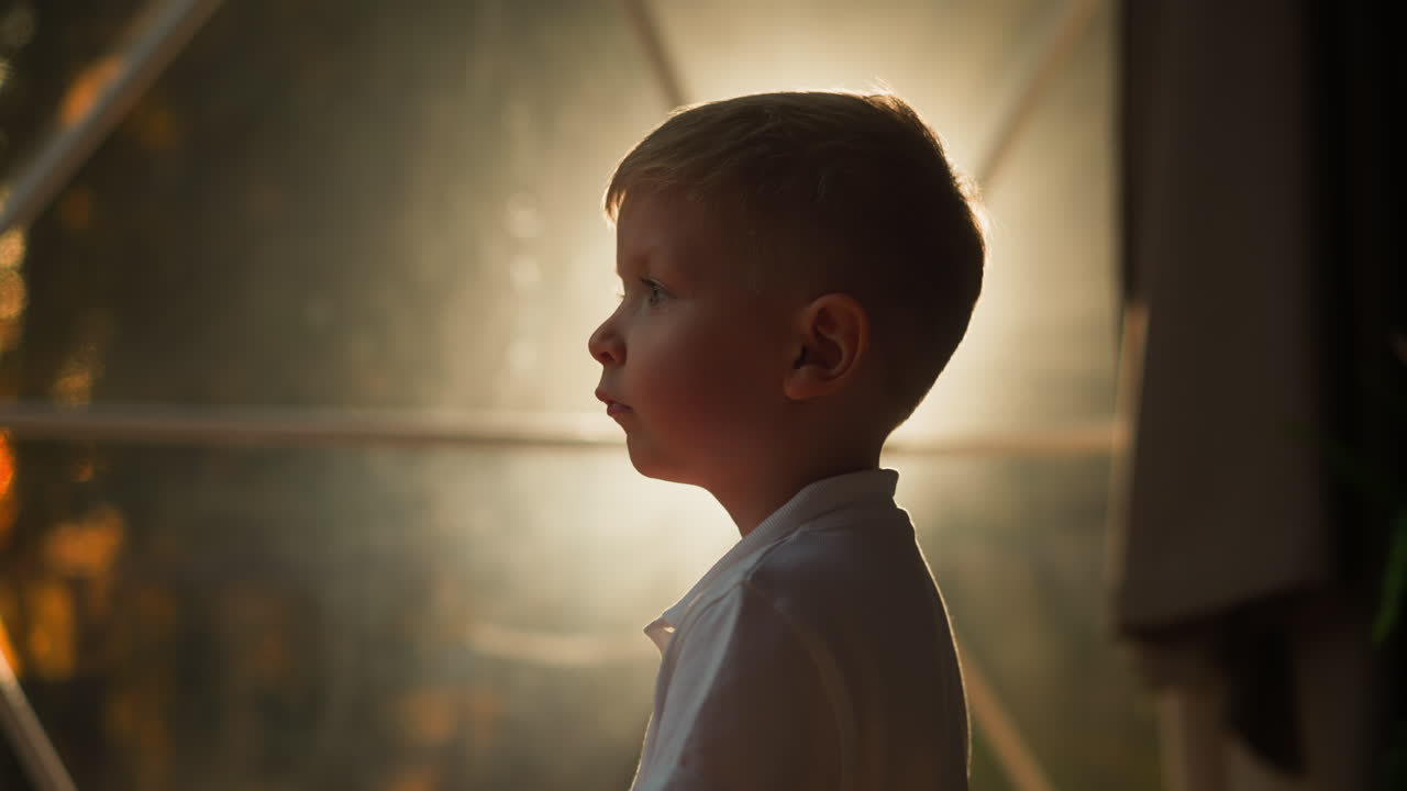 화가 난 아이는 텐트의 빛나는 투명한 벽 근처에 서 있습니다. 피곤한 소년은 저녁에 관광객을 위해 근접한 측면 뷰를 검사합니다. 흐릿한 배경에 글램핑 영역에서 휴식을 취합니다.