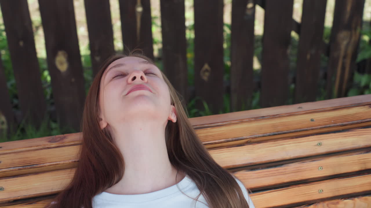 Close up of woman laying back on wooden swing, head tilted upward with closed eyes and peaceful expression, she appears calm and relaxed in backyard setting with wooden fence