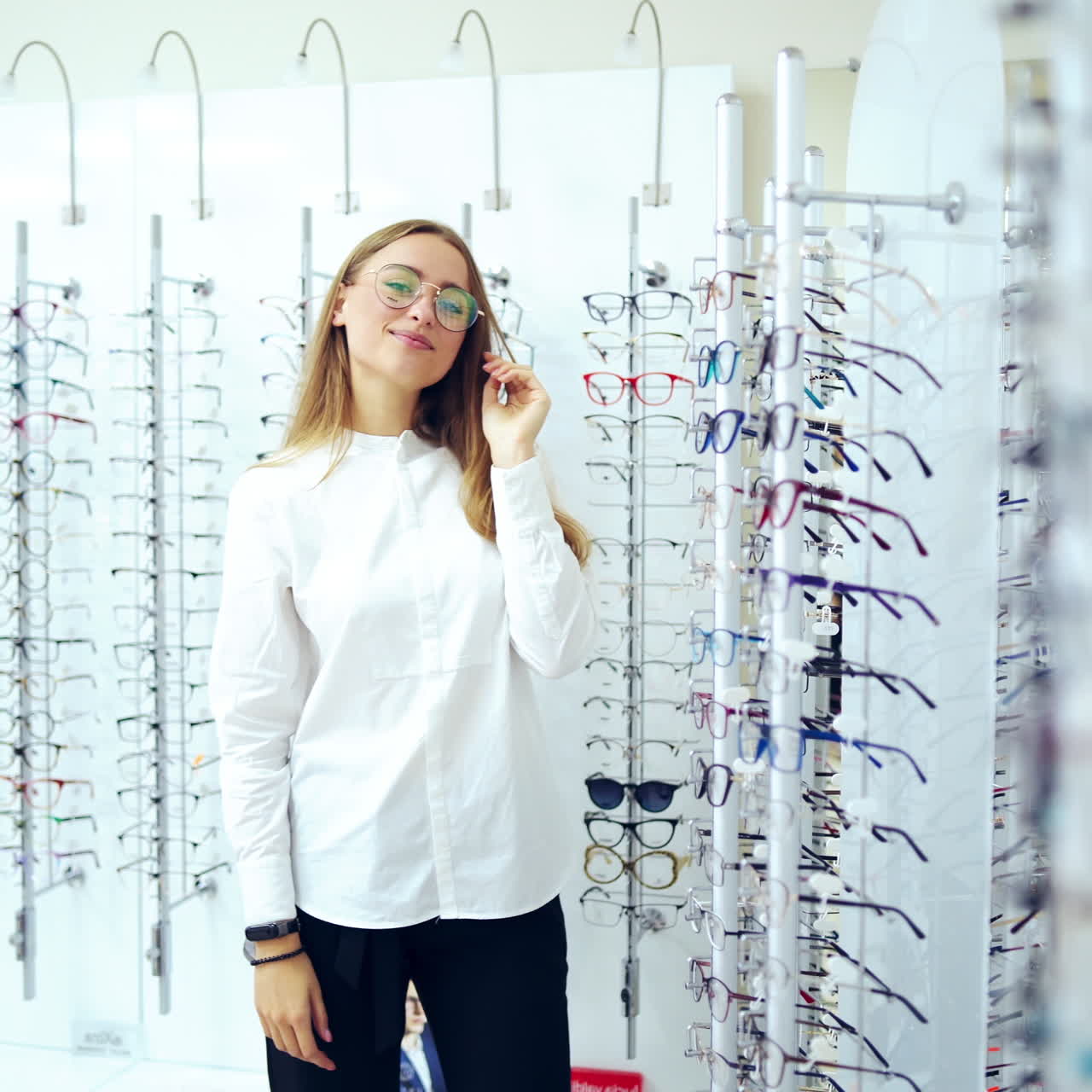 Optical store. Woman choosing spectacles indoors. Young girl in white blouse trying on new eyeglasses on stylish glasses background. Eyesight concept.