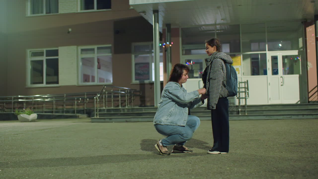 Woman holds hands of young girl in warm jacket outside school entrance early in morning, sharing supportive moment before class, child smiling gently with backpack as streetlights cast soft glow around scene