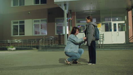 woman holds hands of young girl in warm jacket outside school entrance early in morning, sharing supportive moment before class, child smiling gently with backpack as streetlights cast soft glow around scene
