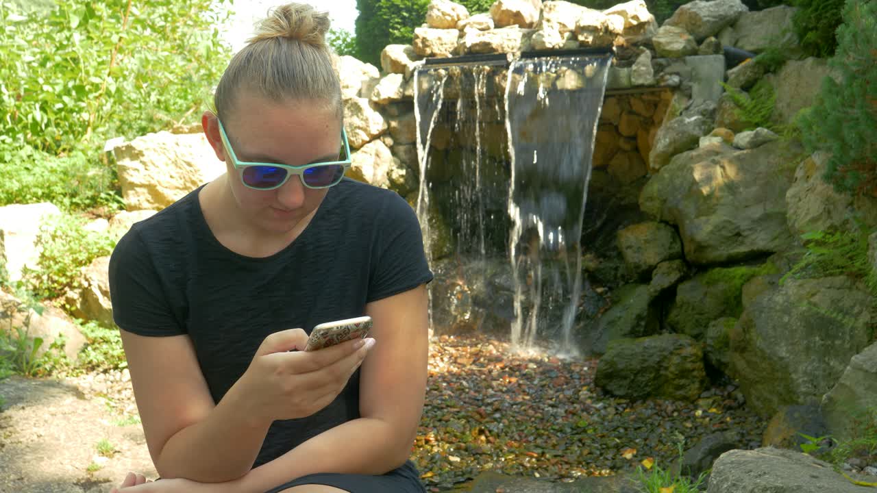 Sexy Woman In Shades Browses Social Media On Her Smartphone While Sitting In A Park Next To A Waterfall