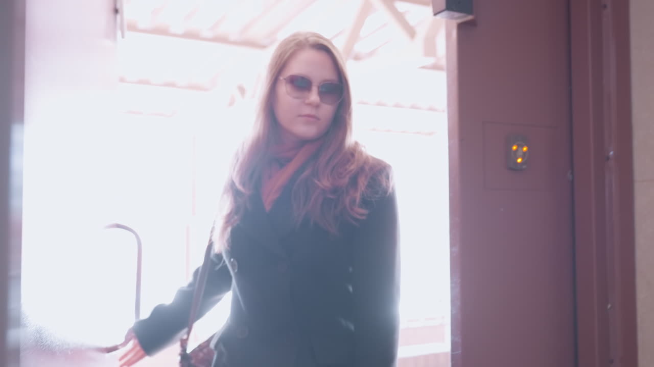 Una elegante mujer con gafas de sol entra por una entrada inteligente, empujando la puerta mientras la luz del sol inunda el pasillo, con un abrigo oscuro y un bolso de hombro que se balancea, con un panel de acceso seguro que brilla en el marco marrón