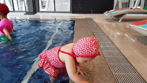 Adorable-Niña-En-Traje-De-Baño-Rojo-Impreso-De-Pie-En-El-Paso-De-La-Piscina-Y-Jugando-Con-Agua-En-La-Piscina