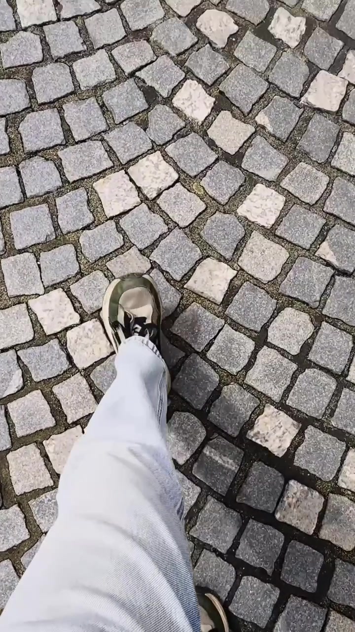 Vertikal förstapersonsvy av någon som går över mönstrad kullerstensbeläggning, som visar en sneaker och vita byxor över en texturerad historisk gatuyta.