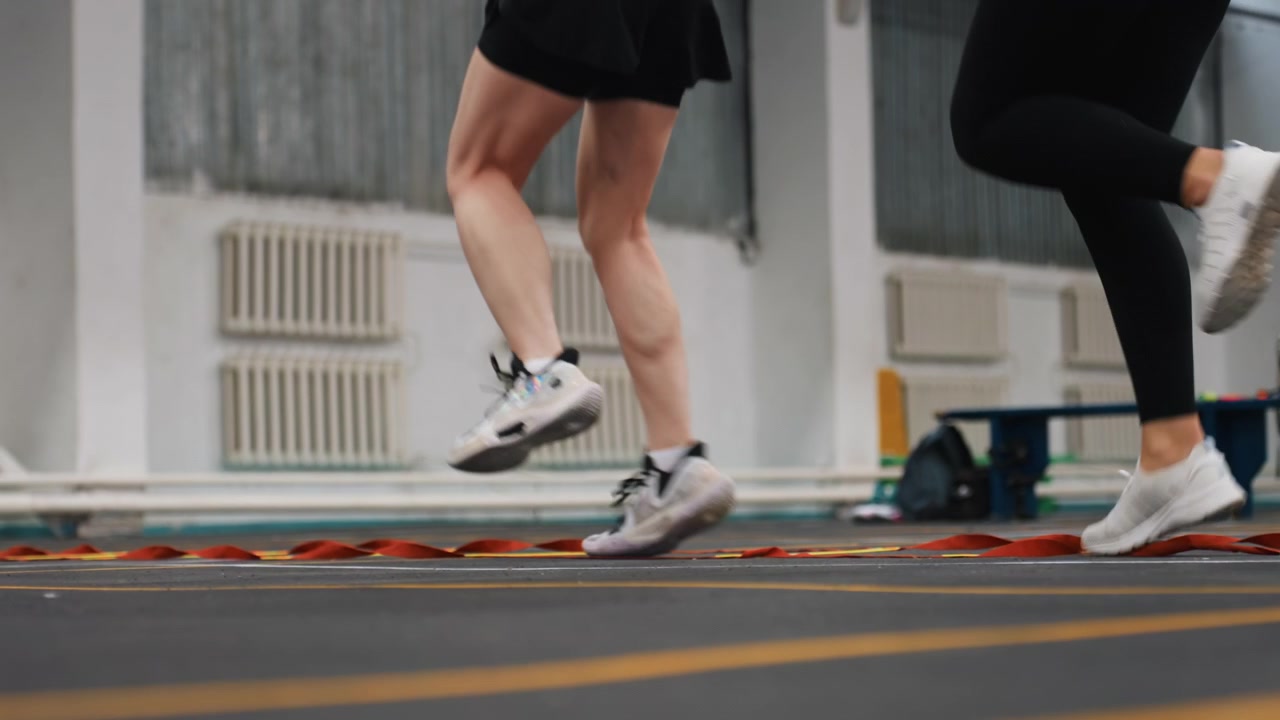 Piedi sul campo inquadrati dal basso che alimentano lo sprint con la scaletta nel badminton, due atleti che eseguono salti esplosivi e scatti rapidi attraverso la scaletta di agilità, movimento dinamico, luci della palestra, impatto delle calzature e intensità pura