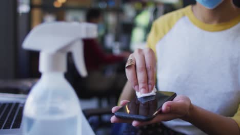 mid section of african american woman wearing face mask cleaning her smartphone using tissue and dis