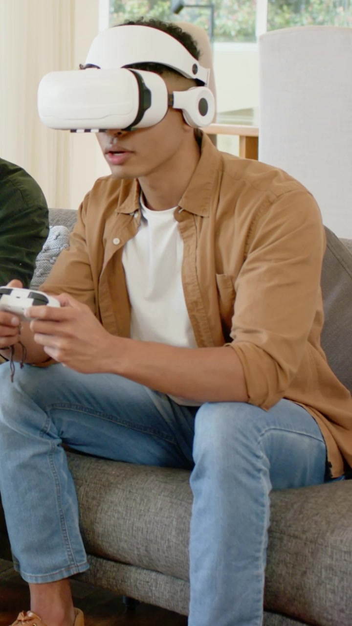Video verticale: Gioco in avvio, un uomo con camicia color cachi e visore VR bianco usa un gamepad cablato in soggiorno.