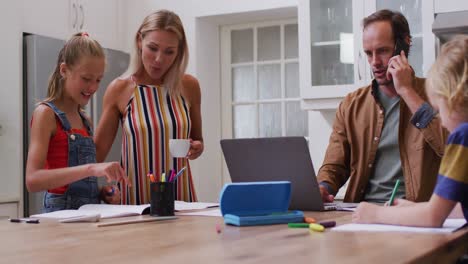 Padre-Caucásico-Usando-Una-Computadora-Portátil-Y-Un-Teléfono-Inteligente-En-La-Cocina,-Esposa-Ayudando-A-Los-Niños-Con-Las-Tareas-Escolares