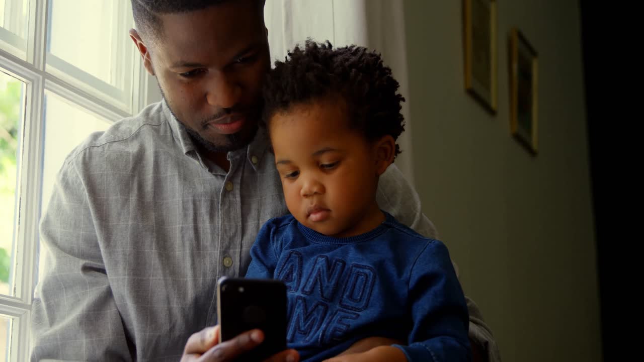 Front view of black father and son using smartphone in a comfortable home 4k