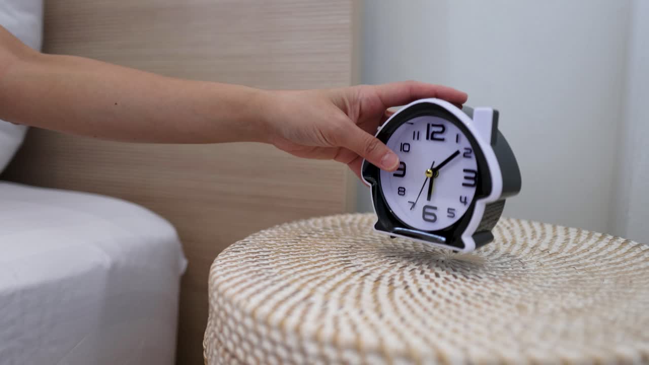 Una mano si allunga dal letto per prendere una sveglia su un comodino di vimini.
