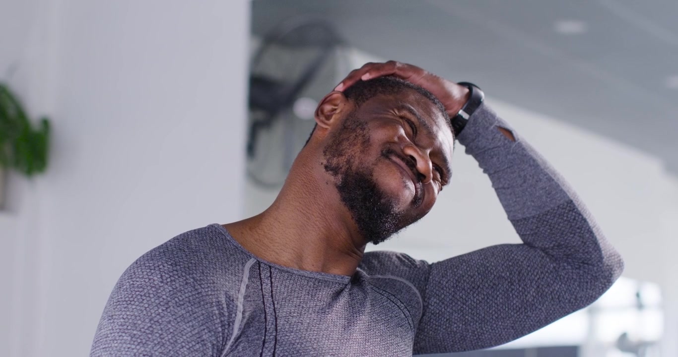 Mid adult African man stretching neck in gym, feeling tension and rubbing scalp with grey top