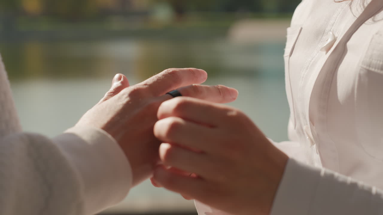 Tender ring placement by caucasian couple. Close framing on fingers and palms beside calm water, soft golden reflections highlight delicate touch and emotional warmth as partners seal vows and deepen