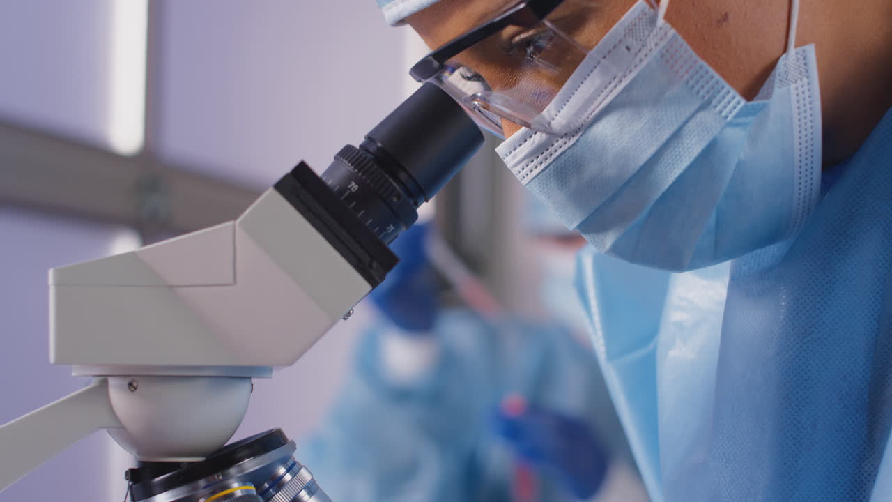 Female Lab Worker Wearing PPE Analysing Samples With Microscope Holding Test Tube Labelled Vaccine