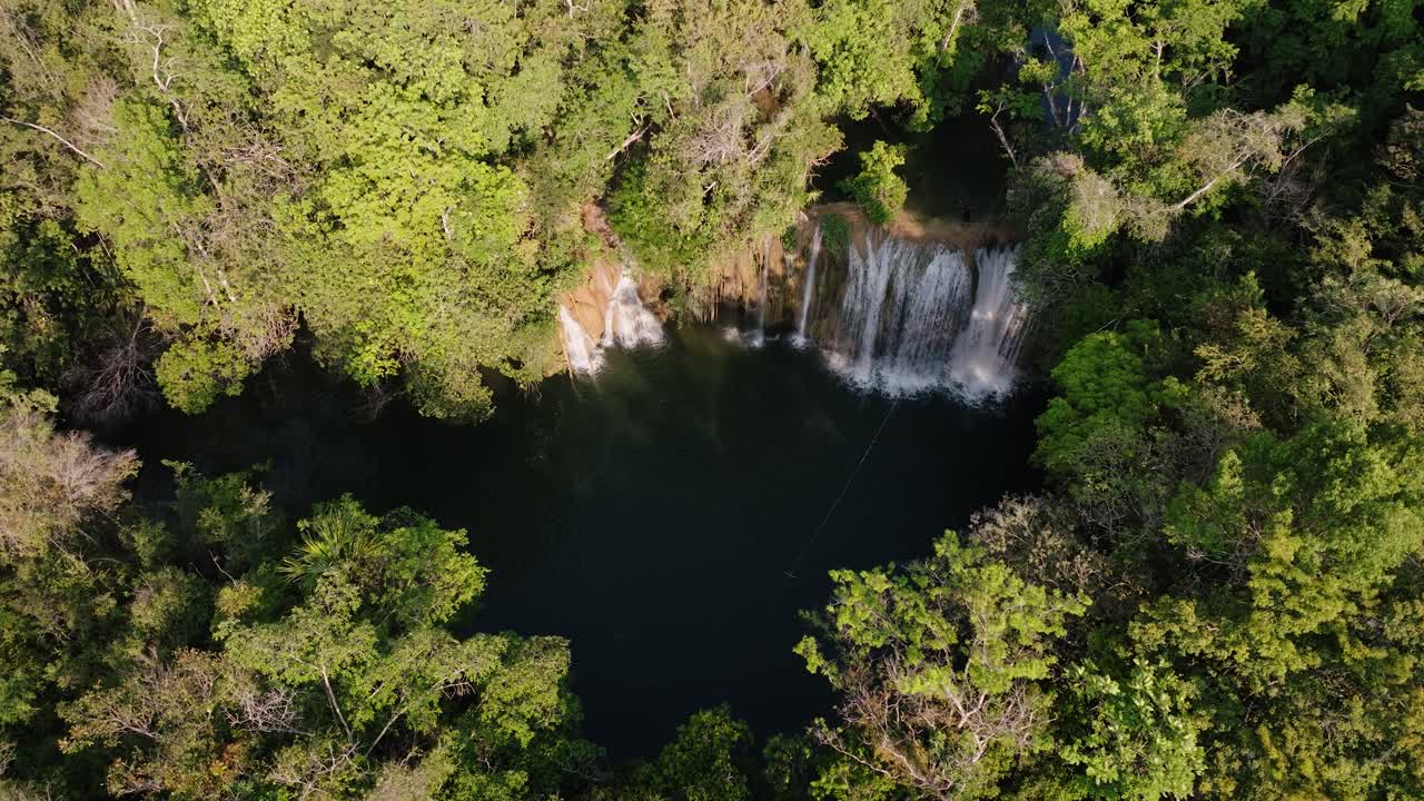 Overhead drone view reveals a secluded jungle waterfall pouring into a calm dark pool surrounded by lush green canopy.