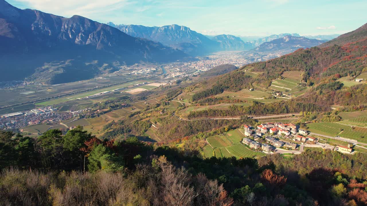 Volo di drone sulla Valle dell'Adige in autunno. Rivela rovine storiche, il fiume turchese, il villaggio di Valsorda con i vigneti e la città di Trento incorniciata dalle Alpi.
