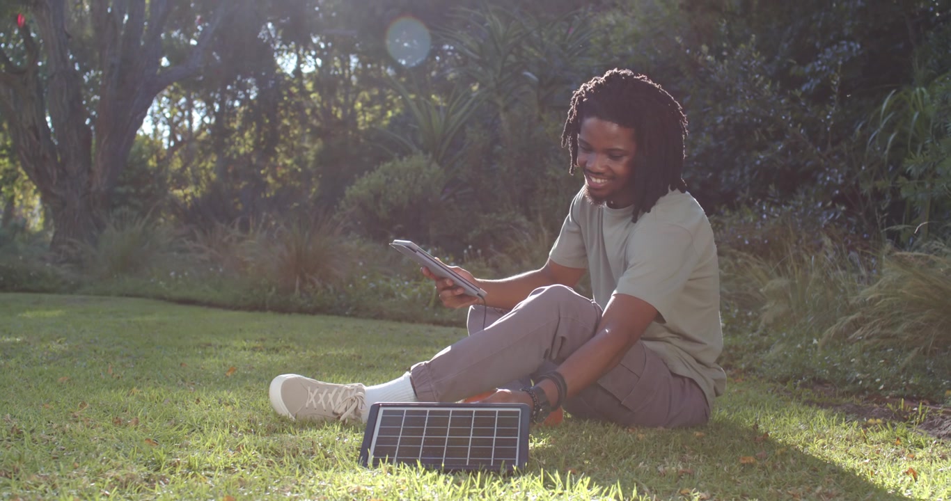 Homme afro-américain inclinant un petit panneau solaire sous le soleil changeant tout en vérifiant la charge de la tablette