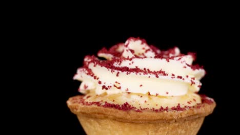 A single red velvet tart with cream cheese frosting rotates smoothly against a solid black background, evenly lit to highlight texture and detail