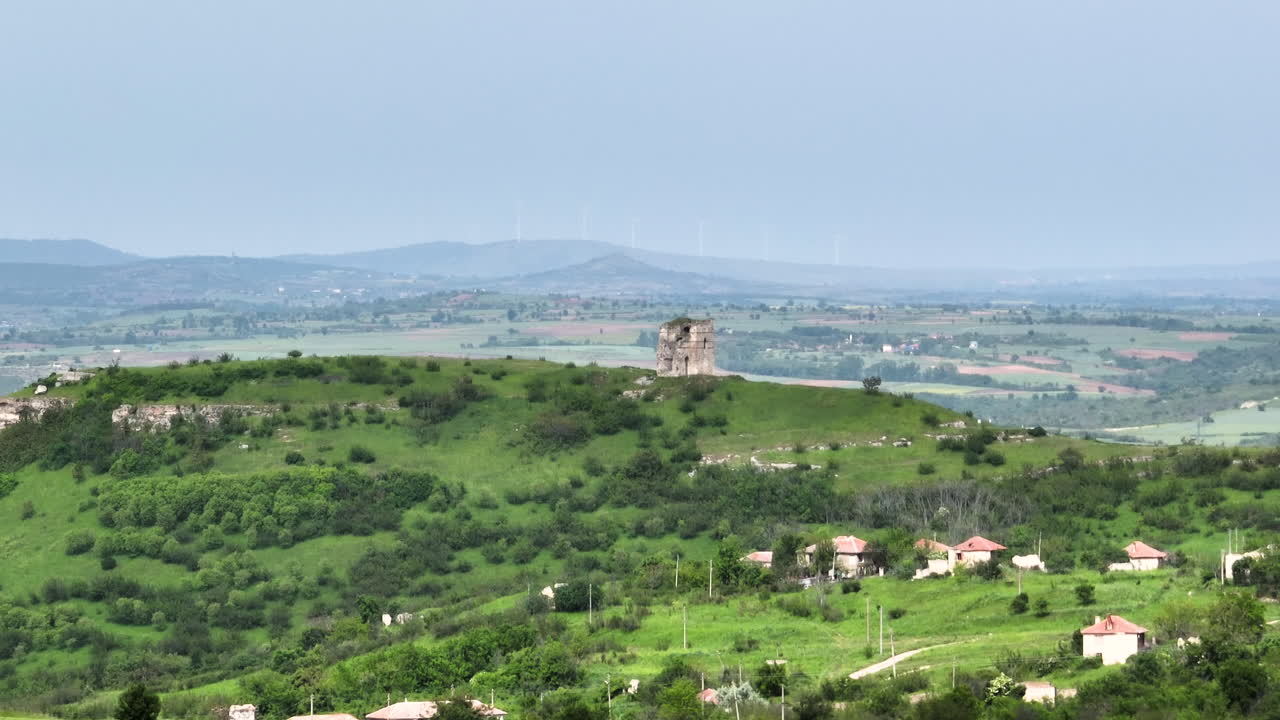 Telephoto parallax drone shot of a hilltop medieval fortress above green rolling hills and village rooftops on an overcast spring day; the access path is visible in the frame