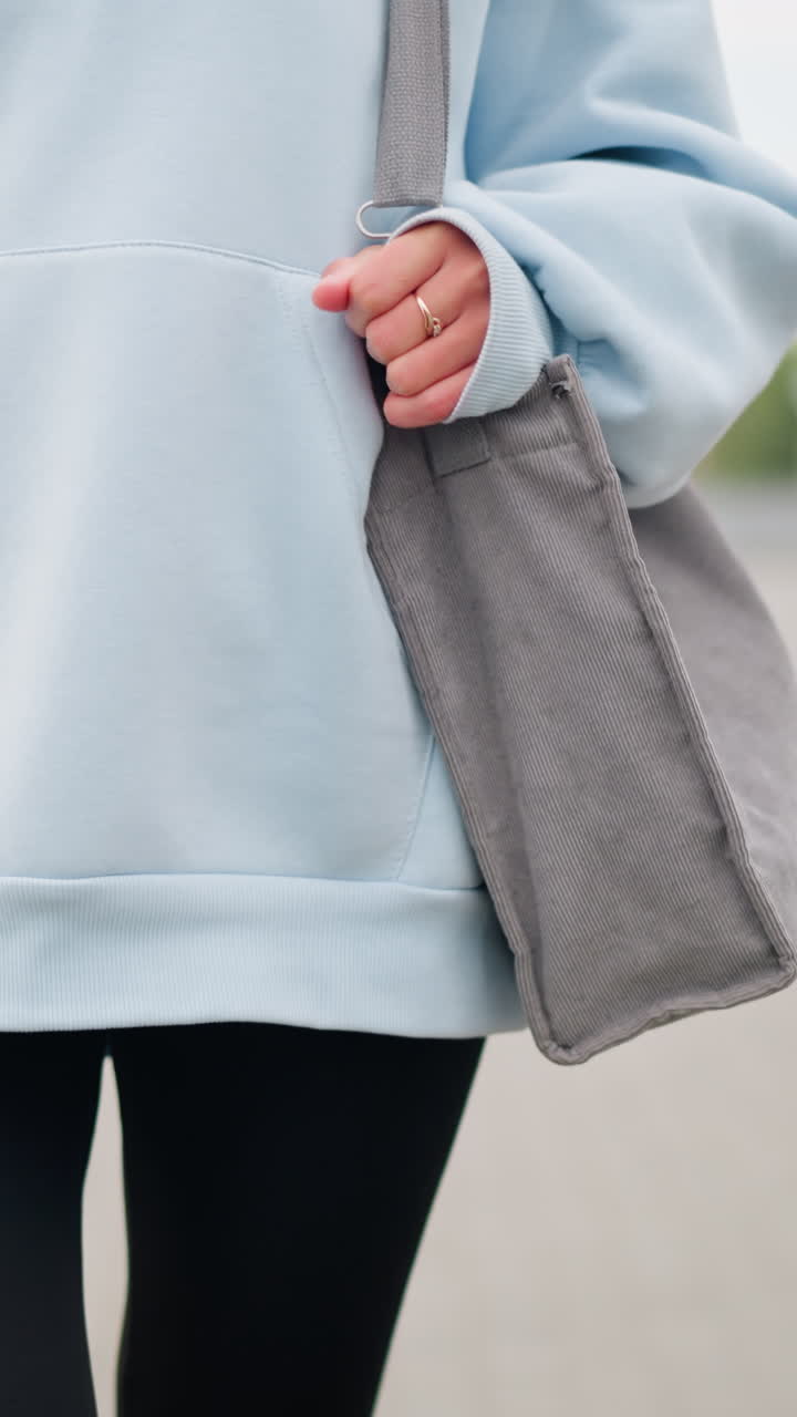 Close-up of young woman in casual attire walking with her bag and golden ring in hand, showcasing her relaxed and confident style, background features a blurred walking path and iron fence