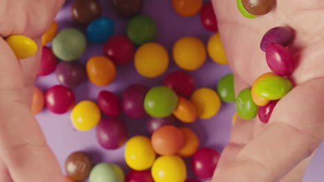 colorful dragee candies in woman hands fall on purple background. summer bright texture studio shot for store, supermarket,close up top view. children sweets made of sugar, mix of lollipops, bonbons.