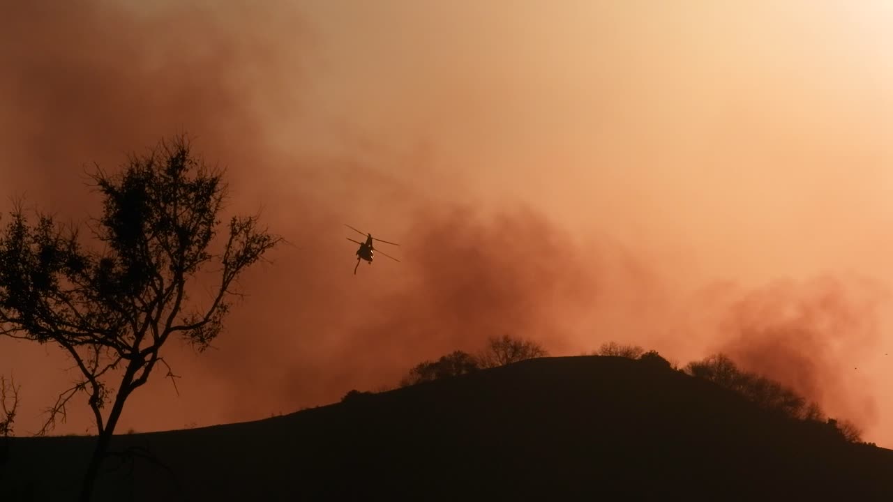 A large wildfire burning in the landscape near Los Angeles, California, with a helicopter flying overhead, likely involved in firefighting efforts. Captures the scale and intensity of the emergency.