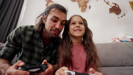 Happy-male-father-in-a-green-checkered-shirt-plays-with-his-daughter,-a-little-brunette-girl-in-a-pink-dress,-using-joysticks,-they-have-fun-and-rejoice-while-sitting-on-a-gray-sofa-at-home-in-a-modern-apartment