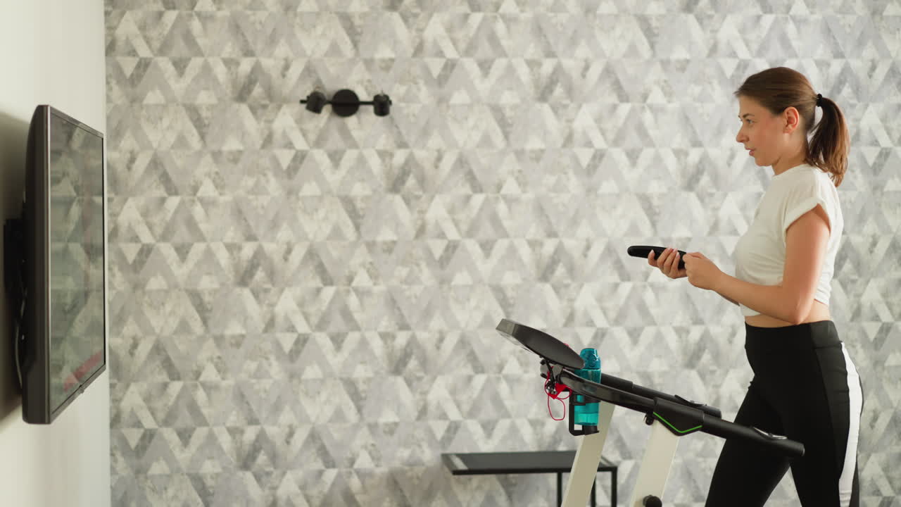 Woman with TV remote control on treadmill at home. Attractive lady turns television channels running on machine in house. Sportswoman amuses on workout