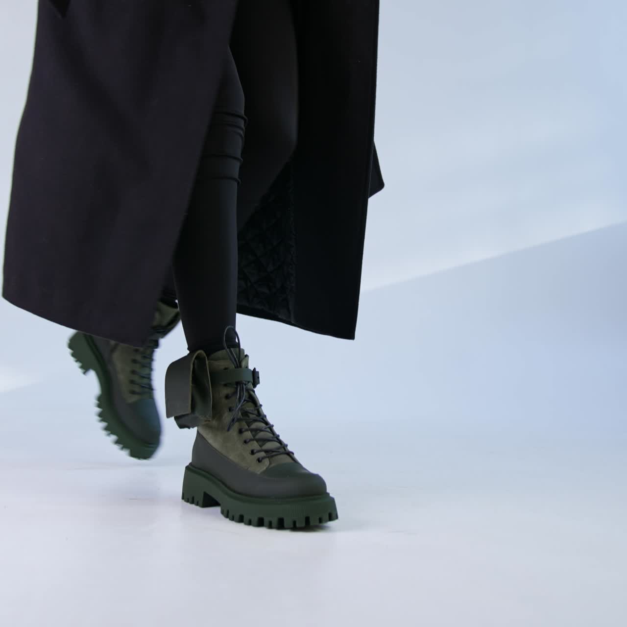 Woman wearing long black coat walk by the studio in her stylish combat boots with little bags on the sides. White backdrop