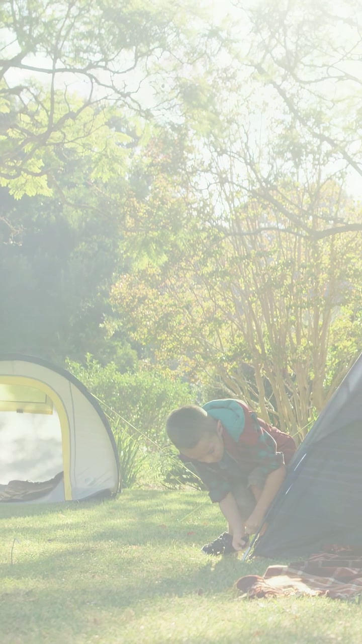 Vertikal video: Barn opdager en løs teltpløk, sidder på hug ved teltet og justerer pløkken, sikrer campingteltet.