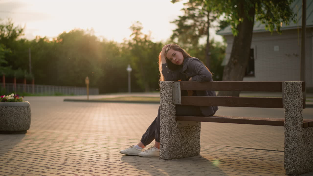 Lady seated at the edge of park bench during golden hour, resting her head on her hand in a thoughtful pose as warm sunlight illuminates the scene, background includes trees, paved walkway