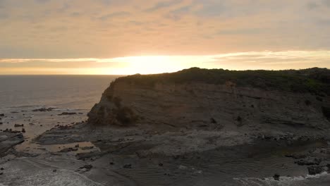 Toma-Aérea-Volando-Hacia-Atrás-Mientras-Mira-Los-Escarpados-Acantilados-En-La-Costa-De-Bass-Victoria-Australia-Con-La-Puesta-De-Sol-En-El-Horizonte