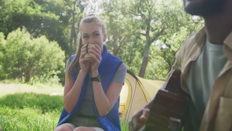 happy diverse couple camping, drinking tea and playing guitar in park, slow motion