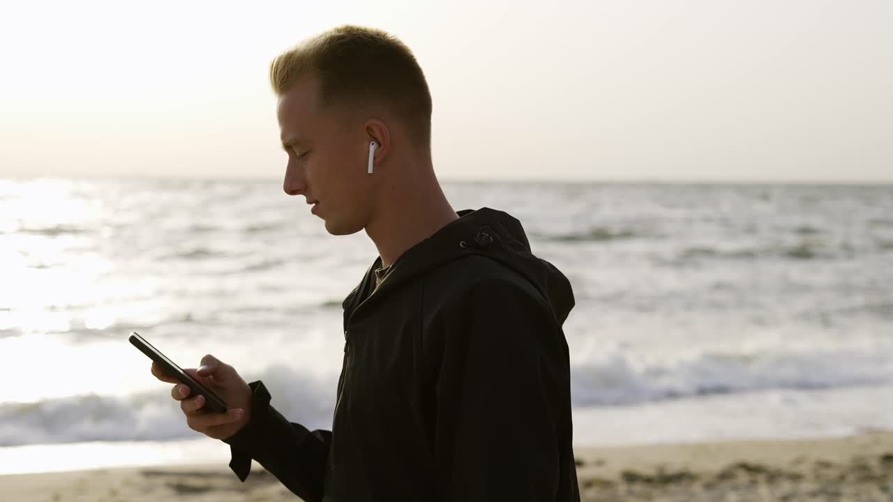 retrato de un joven que escucha música en auriculares usando su teléfono y camina a lo largo de la playa por la mañana