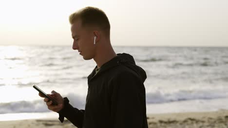 Portrait-of-a-young-man-who-listens-to-music-on-headphones-using-his-phone-and-walks-along-the-beach-in-the-morning